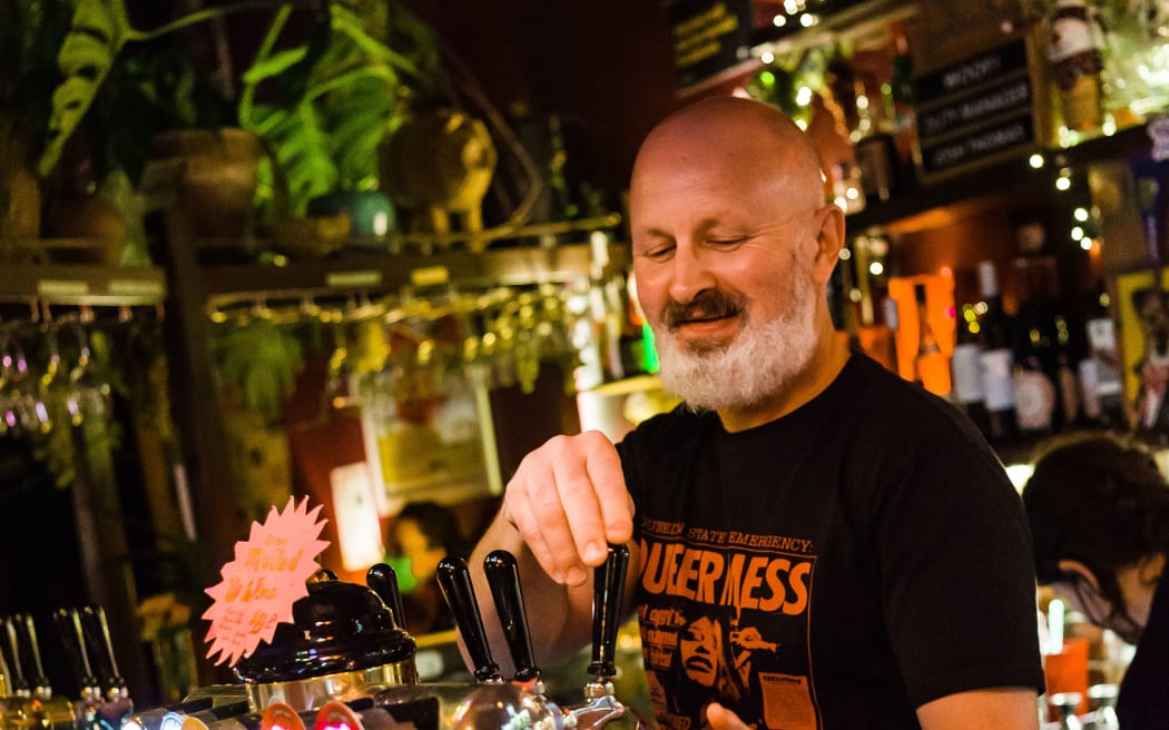 Josh stands behind the bar pouring a beer. He smiles.