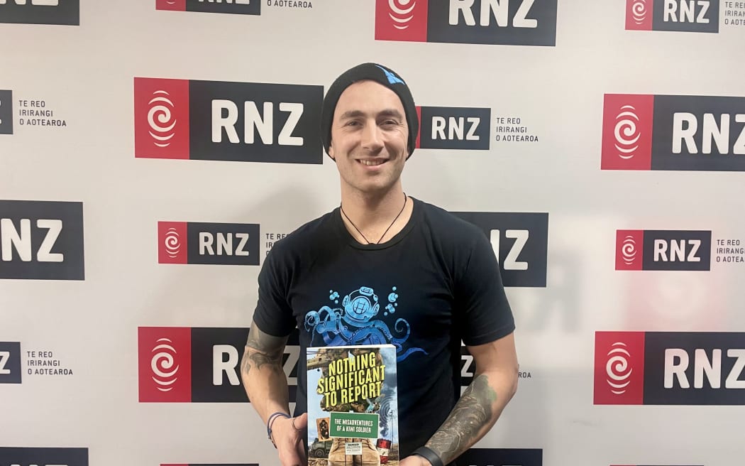 Dario stands in front of an RNZ wall holding his book, 'NOTHING SIGNIFICANT TO REPORT'.