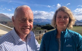 Michael and Meg smile at the camera. Michael wears a navy and white checked shirt, and Meg wears a turquoise outdoors jacket. In the background is Lake Wānaka and snowcapped mountains beneath a blue sky.