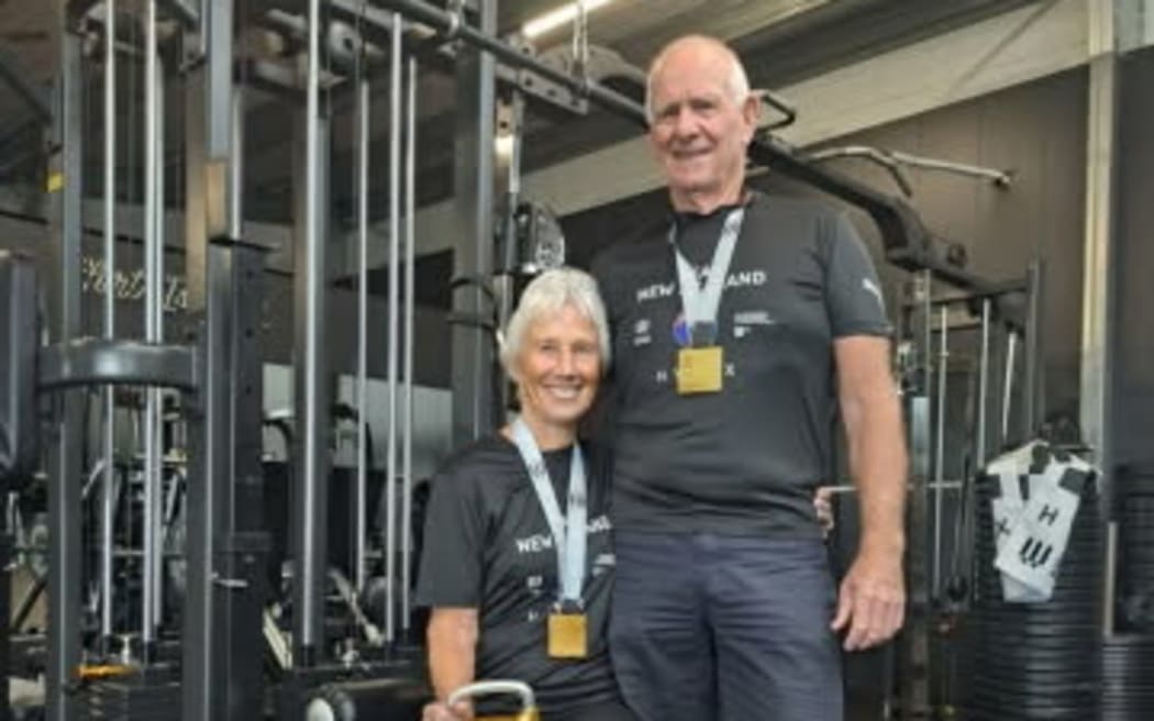 Bob and Sophia Warren stand next to each other in front of some gym equipment. They are wearing gold medals.