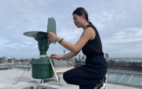 A woman with long dark hair tied back wearing a tank top, long pants and sneakers adjusts a green metal contraption sitting on concrete blocks atop a rooftop with the Sky Tower and Auckland skyline in the background. The sky is cloudy and overcast.