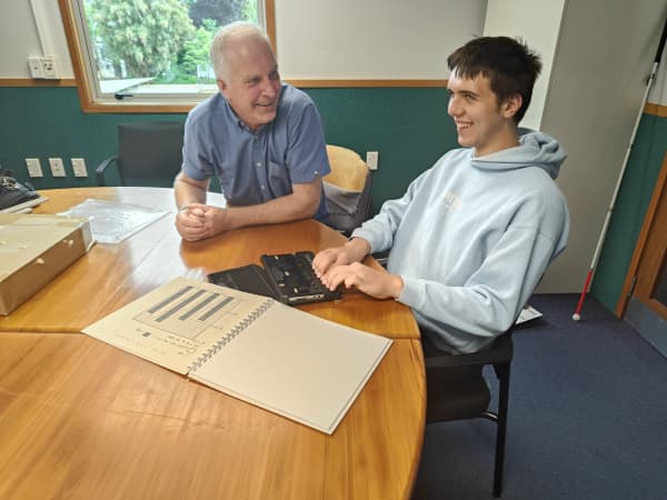 Robin Schofield (left) looks at Toby Ireland (right) as the 17-year-old uses his BrailleSense device on a table with his study booklet.