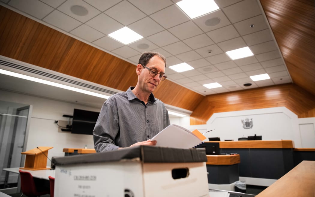 Steven Price reads a file out of a box. He is standing by a desk covered in file boxes in a courthouse.