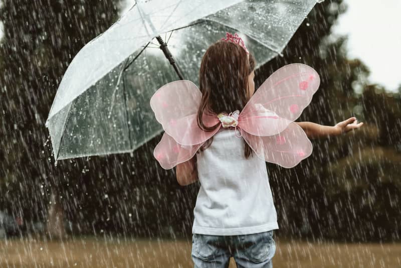 Child in fairy wing costume puts hand in rain under umbrella.