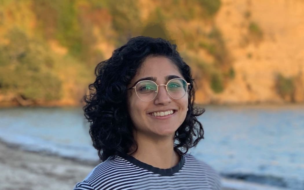 Sachi stands on a beach smiling at the camera. It is evening, and the rocky shoreline behind her is lit up with golden light.
