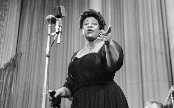 A black-and-white photo of Ella Fitzgerald singing passionately on stage into a vintage microphone, wearing a dark dress and gesturing toward the audience. In Vienna, Austria 23 April 1959.