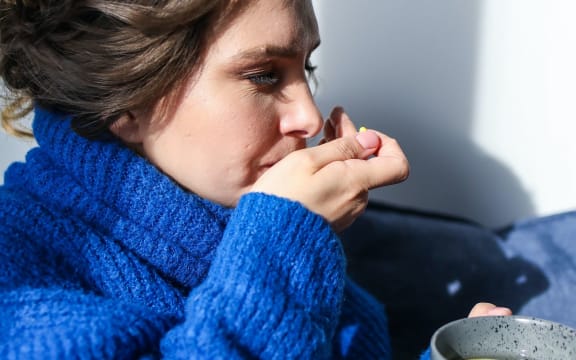 A woman in a blue sweater swallows a pill. She holds a cup of hot lemon water.