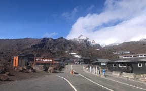 The road into Whakapapa skifield. A wooden sign to the left says "WHAKAPAPA". The sky is a crisp blue. The road disappears up into the snow-capped mountains.