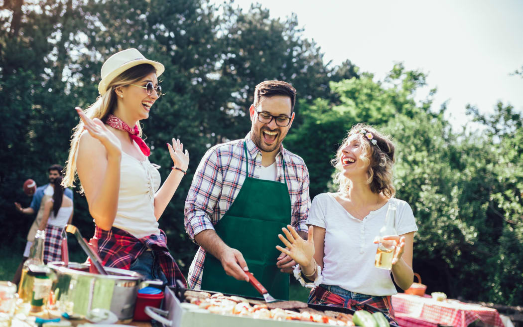 Group of people standing around grill, chatting, drinking and eating.