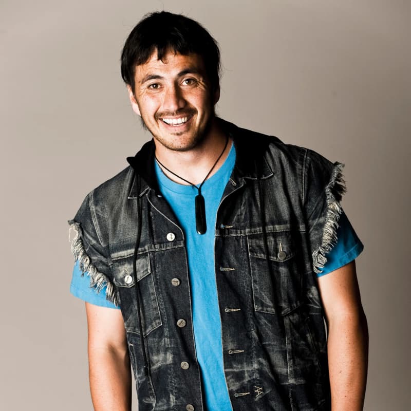 Tammy Davis is a young smiling man wearing a denim vest, blue t-shirt and pounamu pendsnt.