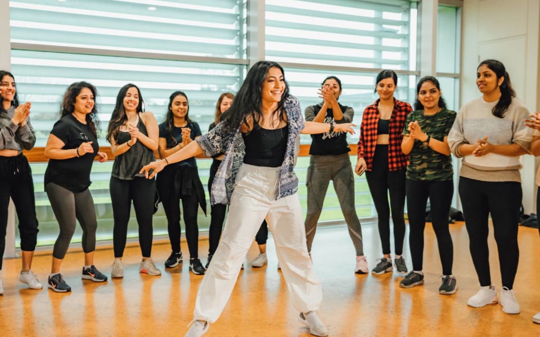 Richa stands in a group of women in a dance studio. She dances and smiles.