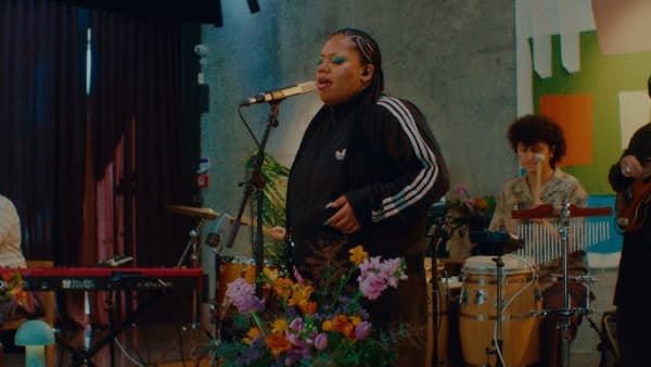 A woman with braided hair, green eyeshadow and a tracksuit top sings with her eyes closed into a microphone.