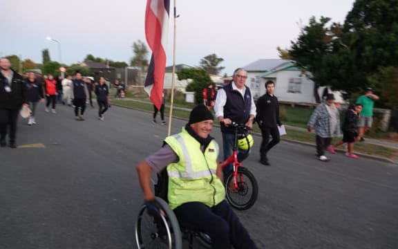 Wairoa resident Val Irwin is in a wheelchair and wears a yellow hi vis vest and black beanie. He has a tino rangatiratanga flag attached to the back of the wheelchair and other people walk alongside and behind him.