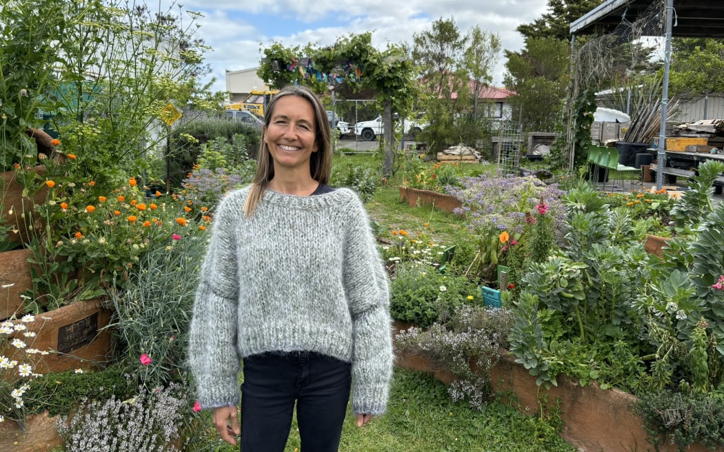 A smiling woman in a woolly grey jumper stands in front of a lush veggie and flower garden.