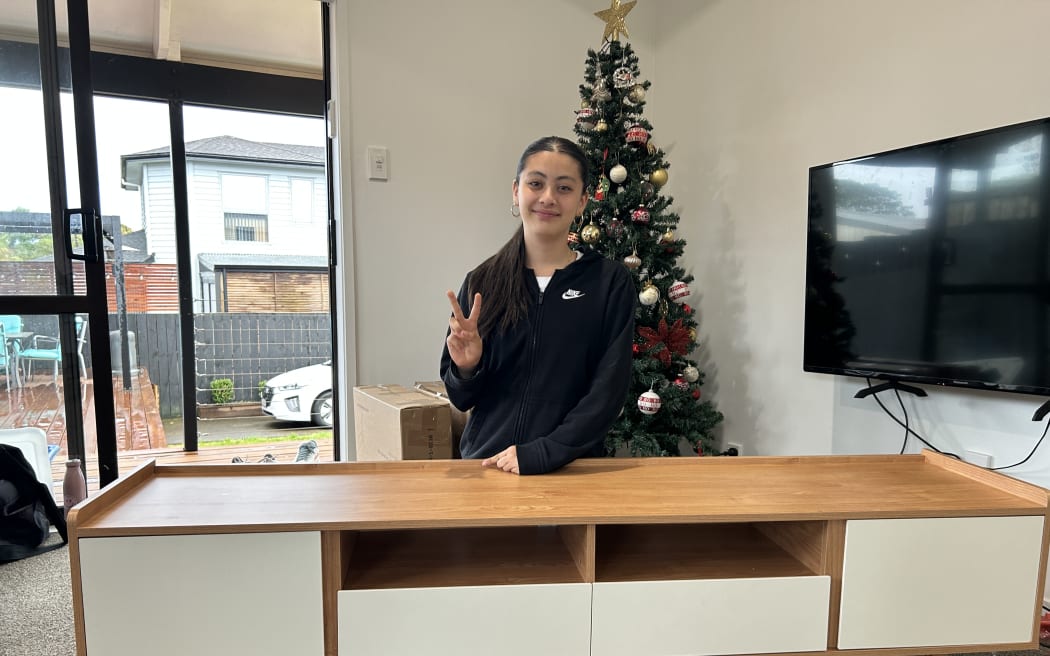 Picture of a young woman smiling at camera, sitting behind an entertainment unit she has just built.