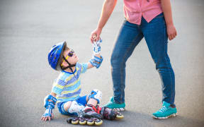 A photo of a Preschooler falling over while rollerblading with mother in the park.