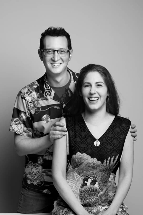 A smiling man with glasses holds the shoulders of a smiling long-harired woman in a sleeveless dress.