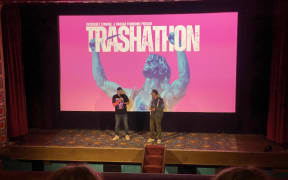 Two men stand at the front of a movie projection screen, introducing an audience before movies play