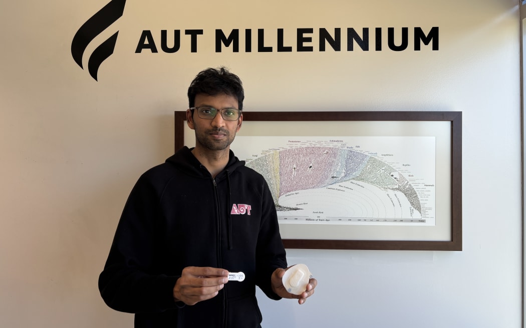 A portrait photo of Anantha against a beige wall, under stencilled black words that say 'AUT Millennium'. Anantha is wearing a Black hoodie, and holding the small sensor in his right hand, and the larger plaster-like sticker to attach it to the participants leg.