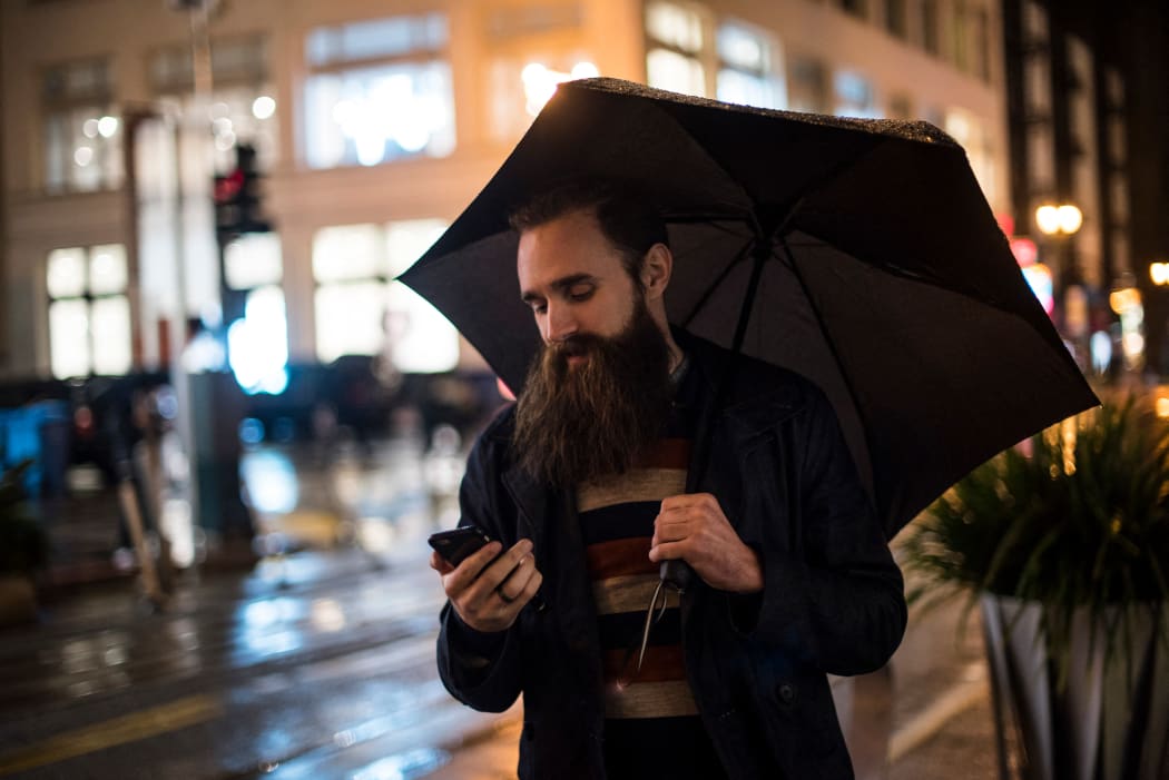 Mid adult man walking in city at night, using umbrella, looking at smartphone, Downtown, San Francisco, California, USA (Photo by Wonwoo Lee / Image Source / Image Source via AFP)