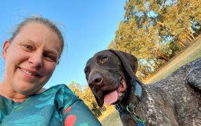 A woman with her blond hair tied back in a ponytail smiles as she takes a selfie with a brown and white speckled pointer. She is wearing a green patterned top. The dog has his tongue hanging out and is wearing a blue collar. There are trees in the background and a clear blue sky.