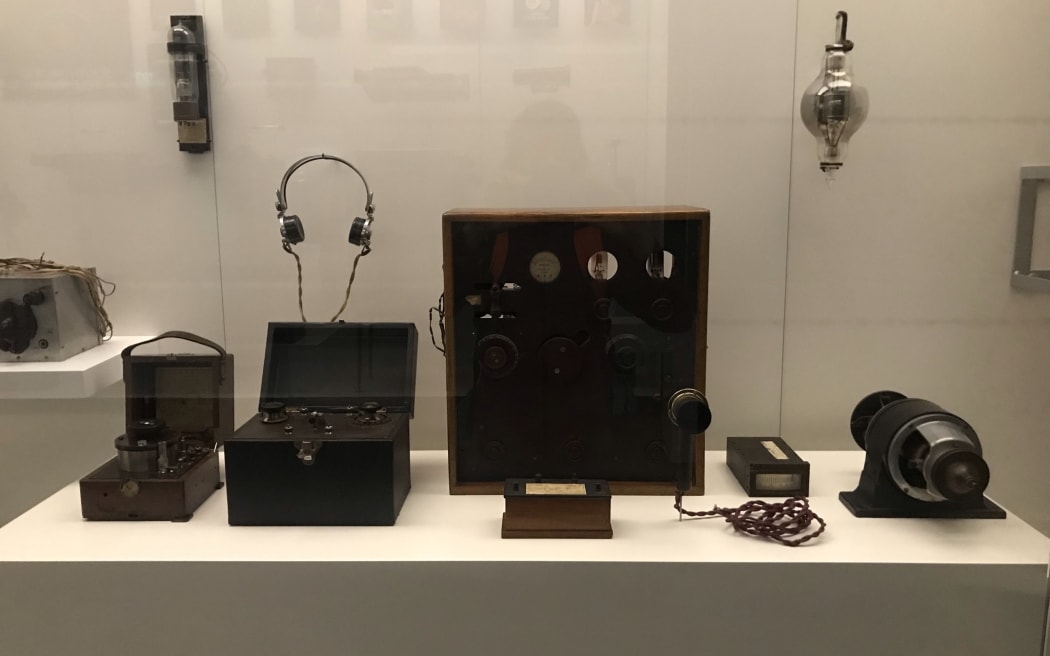 Shows a display case with some early radio equipment used by Professor Robert Jack. A large wooden framed transmitter sits in the centre with a generator and microphone to the side.