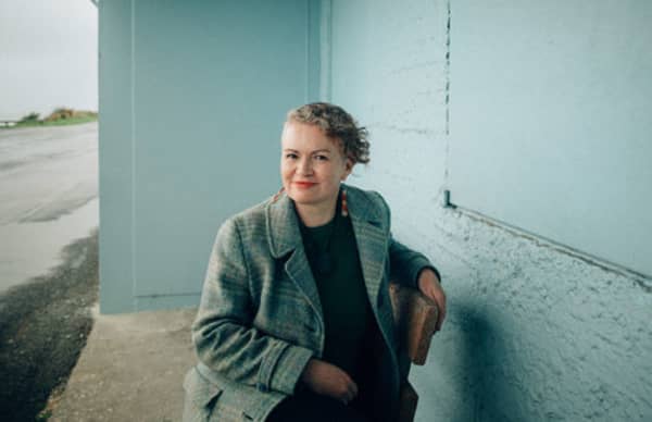 Tina Makereti, a woman in a green coat slightly smiling, sits at an outdoor table.