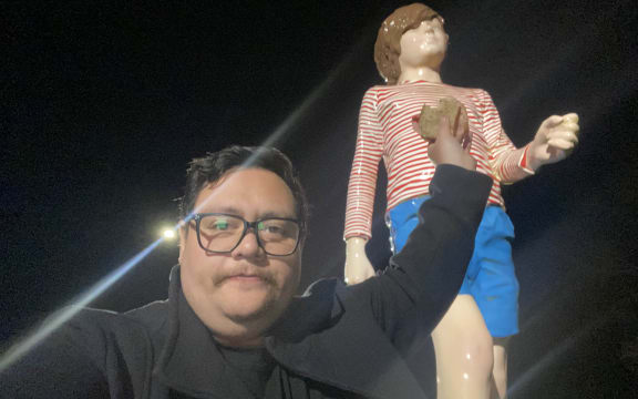 A man in a coat holds up a freshly cooked bacon sandwich up to a giant statue of a kid in blue shorts and a red and white striped shirt.