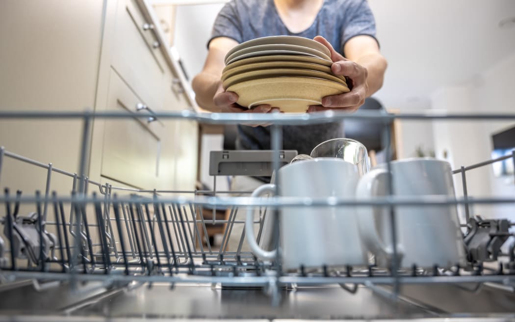 A man in front of an open dishwasher takes out or puts down dishes.
