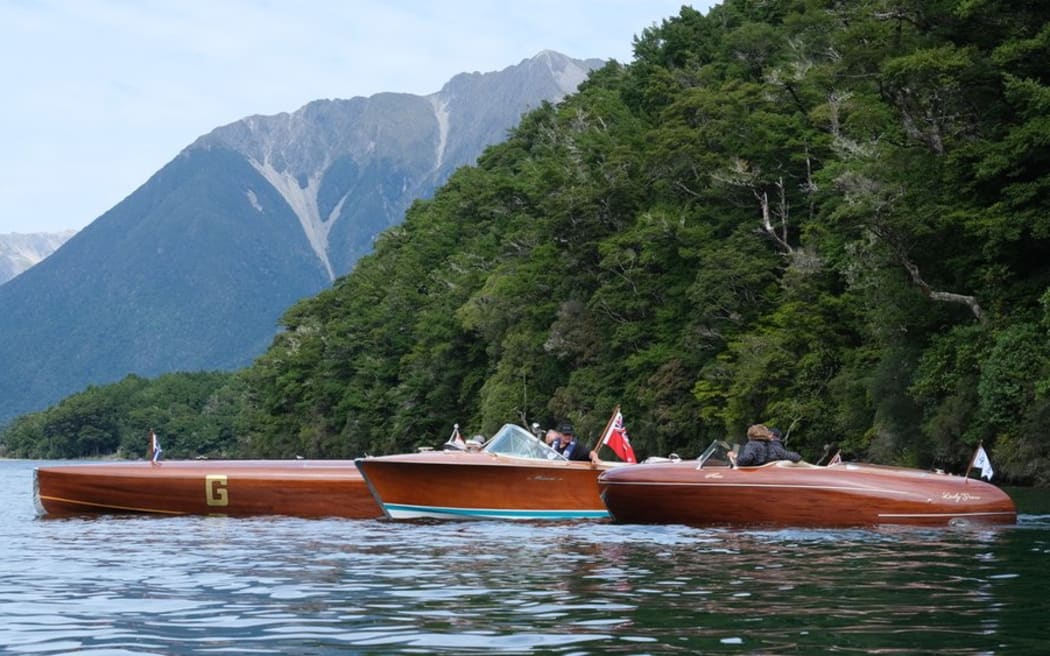 Three wooden boats glide along a glassy lake. Steep, forested mountains rise in the background.