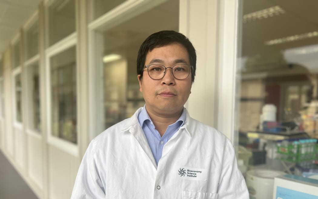 A portrait of Dr Yi Chen in a white lab coat with a Bioeconomy Science Institute logo. He's standing outside a lab, equipment and flasks can be seen through the lab window.