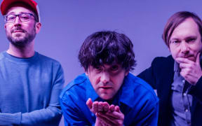 All three members of Bright Eyes lined up in front of a blue background.