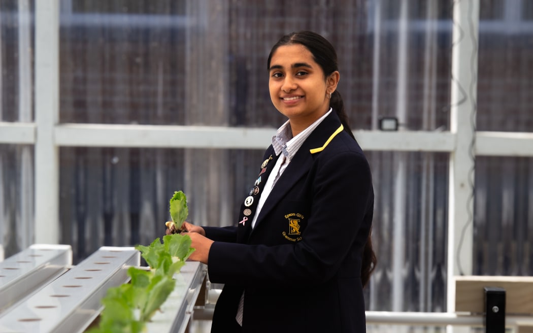 Rena is in her school blazer, looking at the camera and smiling. She is standing in front of a tray of lettuce plants, holding one in her hand.