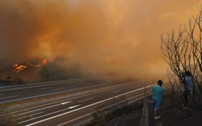 A wildfire burns near a highway in A Gudina, northwestern Spain, on August 15, 2025. All of Spain was on a heatwave alert today, while the weather agency warned that much of the country was at "very high to extreme risk" from wildfires. Nearly two weeks of high temperatures have left Spain sweltering, and today spread to Cantabria, which has so far been spared from the searing heat. Temperatures in the northwestern region were forecast to pass 40C, the national meteorological agency, Aemet, said. Spain has endured a devastating season of fires, with 157,501 hectares (389,193 acres) reduced to ashes since the start of the year, according to data from the European Forest Fire Information System (EFFIS). (Photo by CESAR MANSO / AFP)