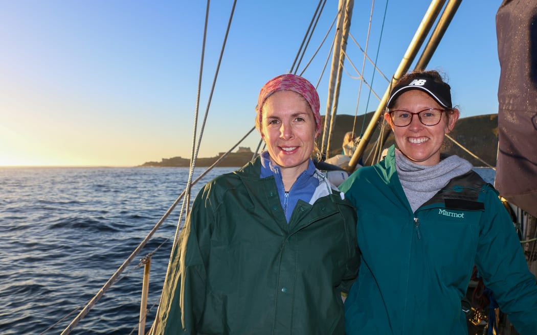 Two women wearing warm rainjackets stand on the deck of a yacht on the ocean. On the left the woman's jacket is forest green and she has a warm headband on. The woman on the right has a teal jacket, glasses and a visor cap. Behind them are the steep cliffs of the main Antipodes Island silhouetted against the fading twilight.