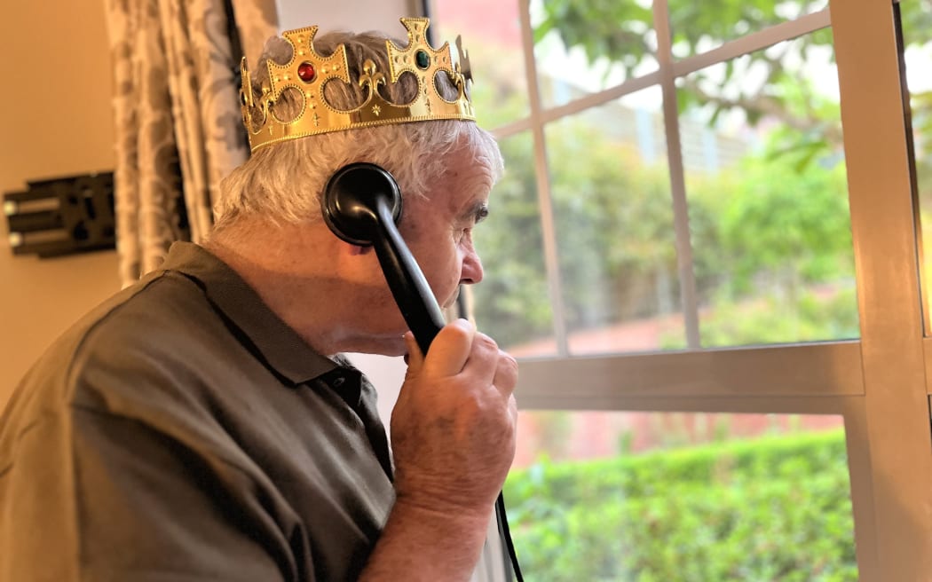 Tom, an elderly man in a brown t-shirt, looks out a window wearing a golden crown and holding an old-fashioned black rotary phone.