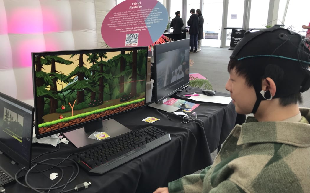 A young student is sitting in front of a computer with an EEG cap on looking at a screen with an angry birds look-a-like game on it.