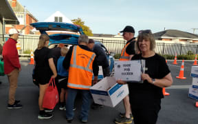 A woman dressed in black called Minnie is looking at the camera and holding a sign that says "Toot for tucker". Behind her a few people with their backs to the camera are loading boxes of donated food into the back of a car.