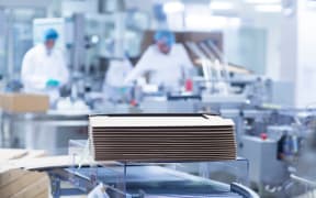 Workers on production line in apharmaceutical plant, cardboard boxes stacked in foreground.