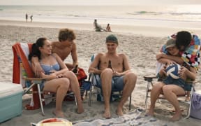 Five young people relax on a beach in deck chairs.