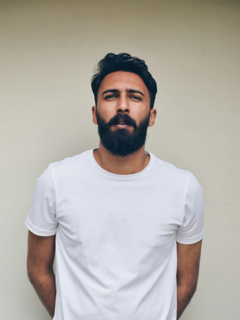 A bearded and brown-skinned and bearded man wears a crisp white t-shirt and stands against a beige wall.