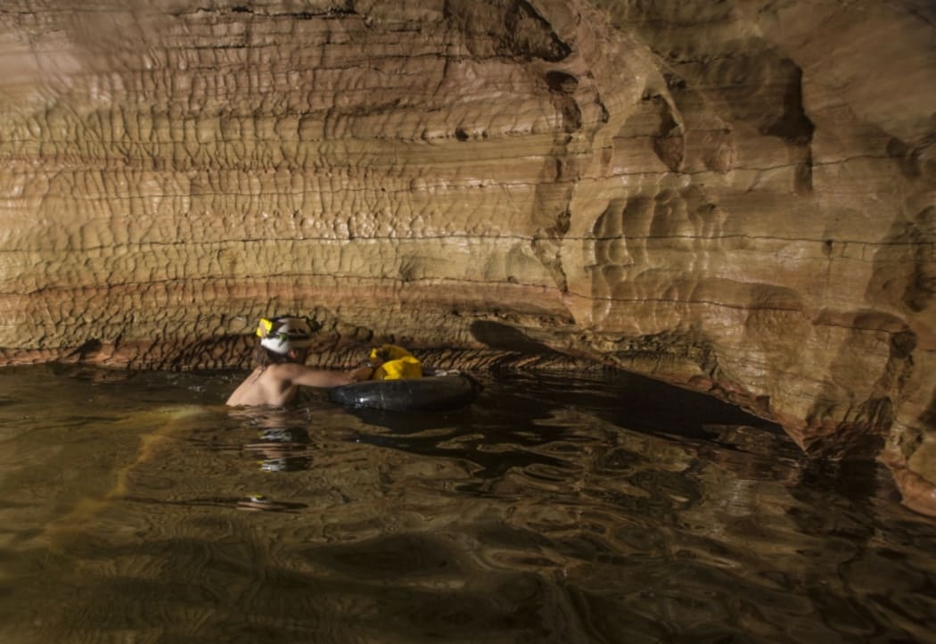 An image of a caver swimming across a freezing cold stretch of water inside Armageddon cave. Their equipment is  inside a dry bag.