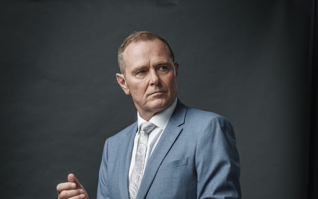 Actor Stephen Lovatt stands dressed in blue suit against a soft grey background.