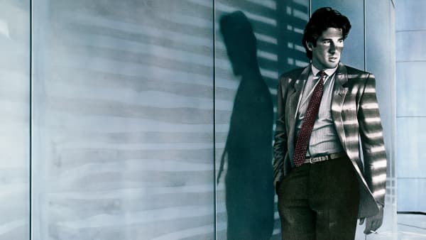 Richard Gere - an attractive man in a suit jacket and tie - stands in moody lighting.