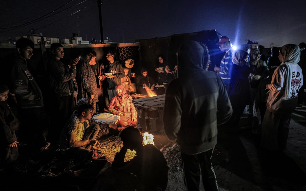 People gather around women preparing meals by a stove outdoors at a camp for displaced Palestinians in Rafah in the southern Gaza Strip near the border with Egypt on 31 December, 2023 amid the ongoing conflict between Israel and the militant group Hamas.