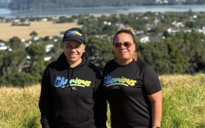 Louisa Tipene Opetaia and Ama Mosese stand atop Te Pane o Mataoho - Māngere Mountain. In the background you can see some houses and the sea.