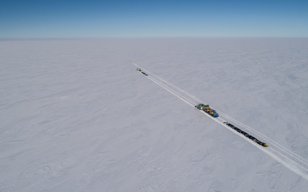 An aerial image looking down on a flat white expanse of ice with three vehicles towing long trailers of gear and small portable buildings. The front vehicle is carving a track parallel to the other two; both tracks extend from the bottom right corner into the middle of the white expanse.