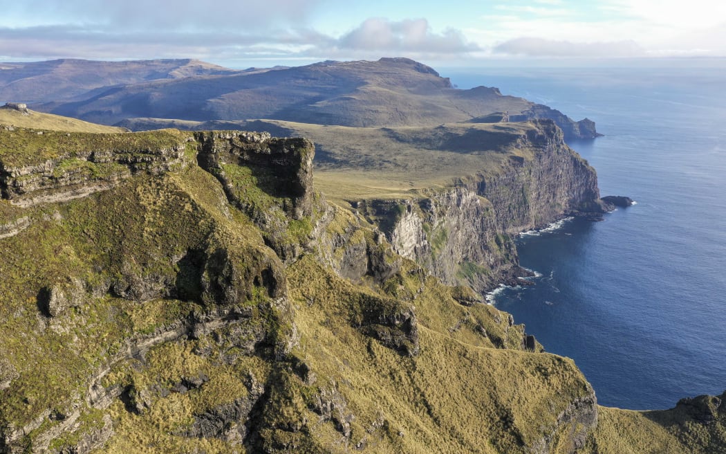Rugged and steep cliffs covered with low, grassy vegetation next to deep blue sea.