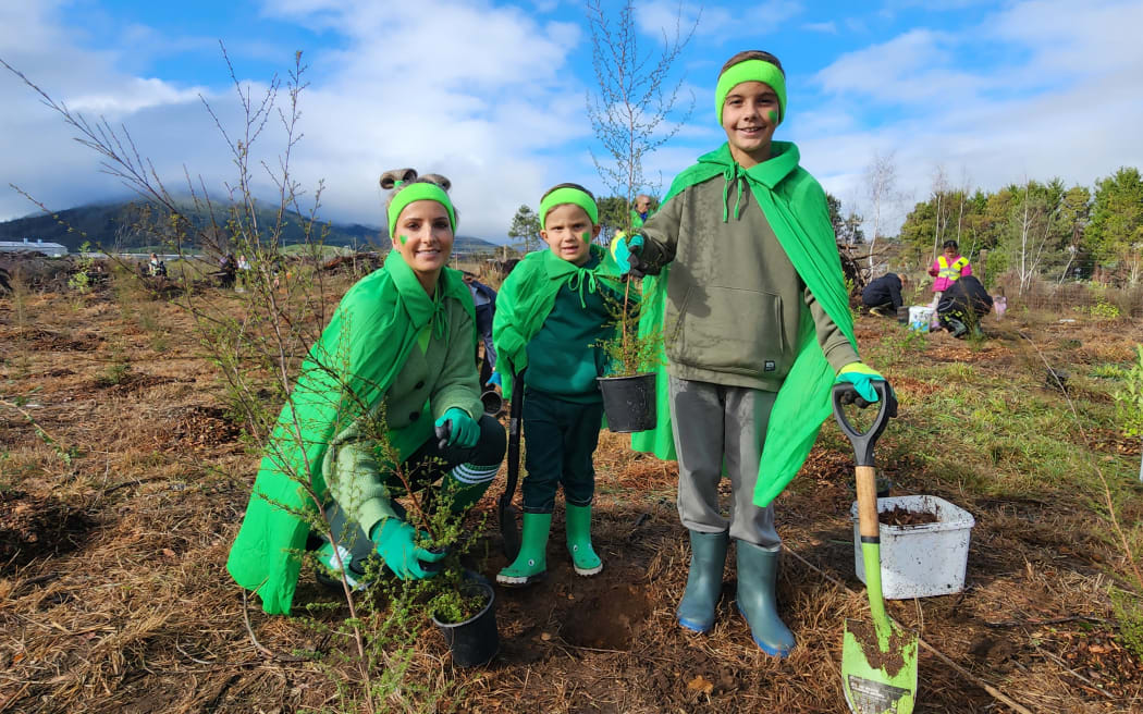 A woman and two children dressed in bright green capes, gumboots and green headbands hold seedlings and a spade at a planting site.