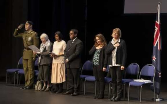 Dignitaries lined up on stage including saluting solider and others with hand over heart.